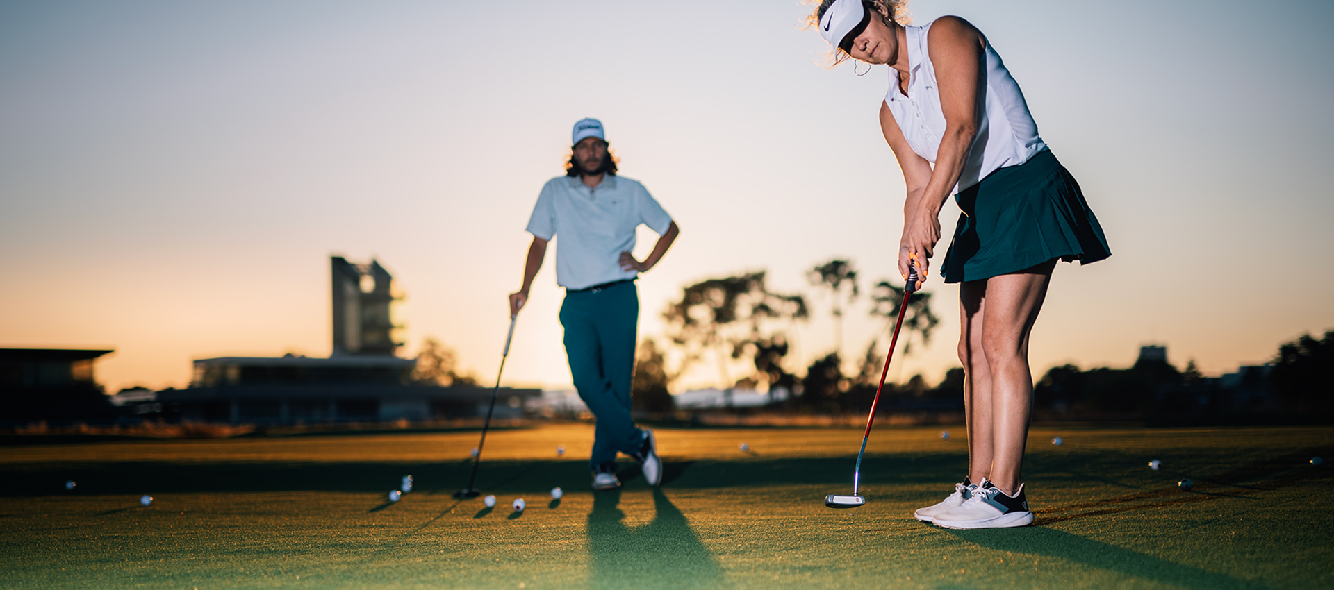 Une femme fait du golf, un homme la regarde. Le soleil se couche au loin.