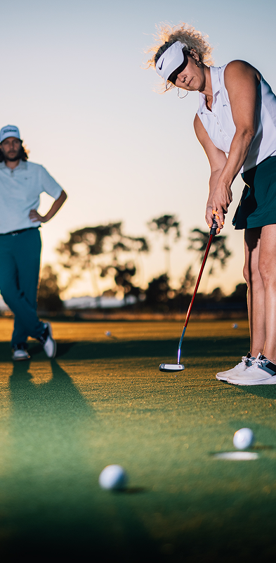 Une femme fait du golf, un homme la regarde. Le soleil se couche au loin.